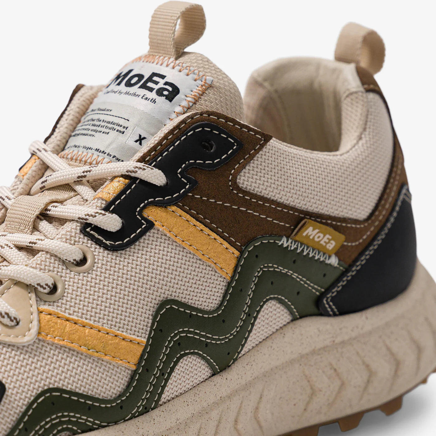 Nahaufnahme eines beigen Sneakers mit braunen, grünen, gelben und schwarzen Akzenten, strukturierten Nähten, einer dicken Sohle und einem Etikett auf der Zunge mit der Aufschrift MoEa. Das Design weist gewellte farbige Streifen an der Seite auf.