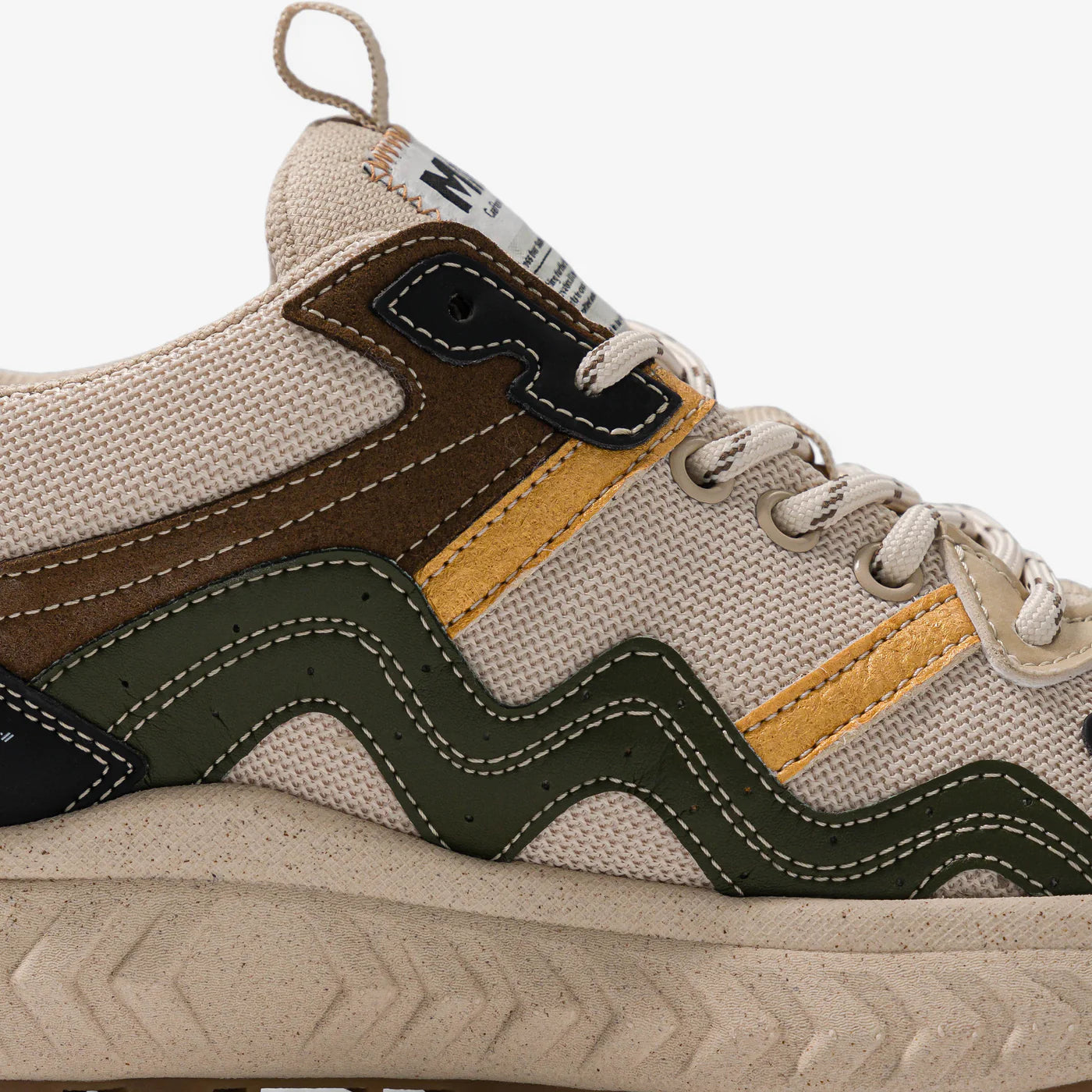 Nahaufnahme eines beigen Sneakers mit strukturiertem Stoff, olivgrünen und gelben Wellenstreifen-Akzenten, braunen und schwarzen Details, beigen Schnürsenkeln und einer gesprenkelten beigen Sohle.