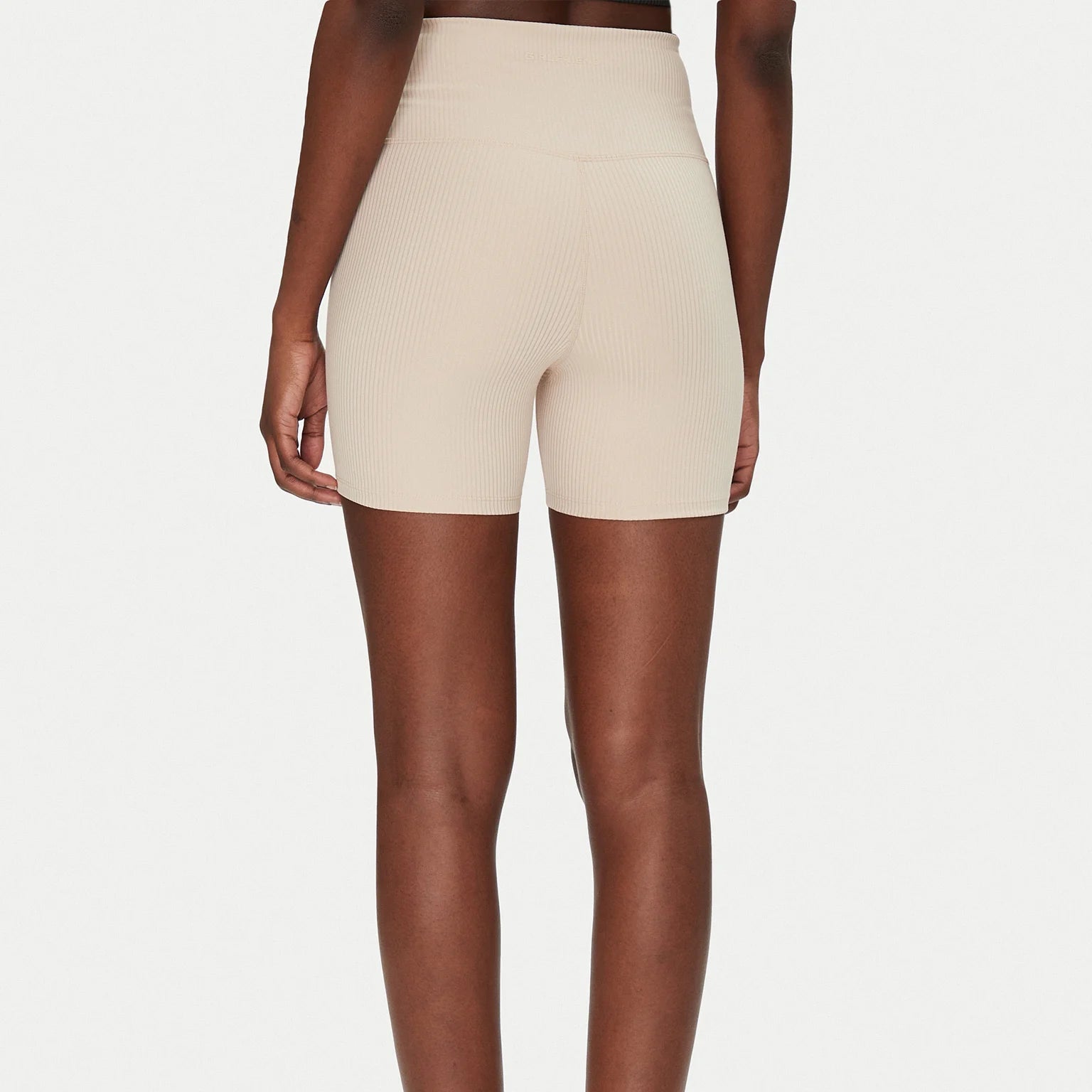 Eine Person steht mit dem Gesicht zur Kamera, trägt eine hochtaillierte, hellbeige gerippte Radlerhose und weiße Turnschuhe. Der Hintergrund ist einfarbig weiß.