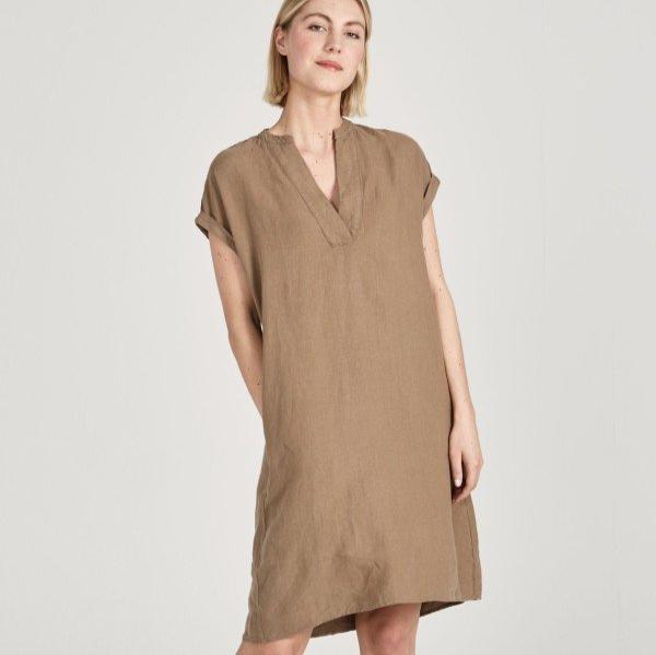 Frau in einem hellbraunen, ärmellosen Kleid vor neutralem Hintergrund.