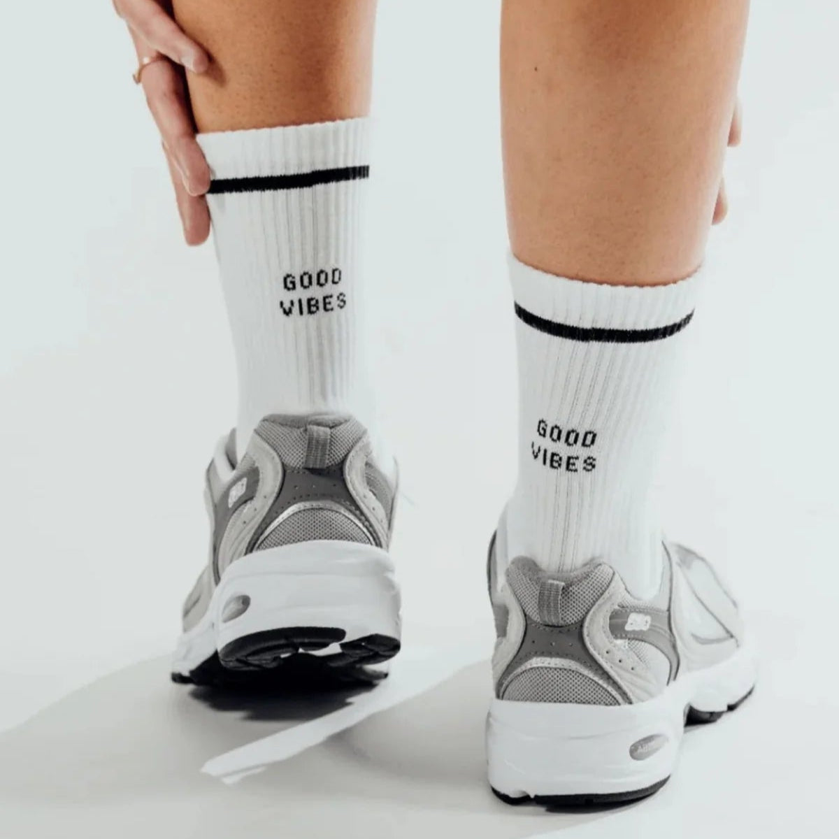 Person in weißen Socken mit der Aufschrift GOOD VIBES und grauen Turnschuhen, die auf einem weißen Boden steht. Eine Hand zieht eine Socke hoch, und nur ihre Beine sind zu sehen.