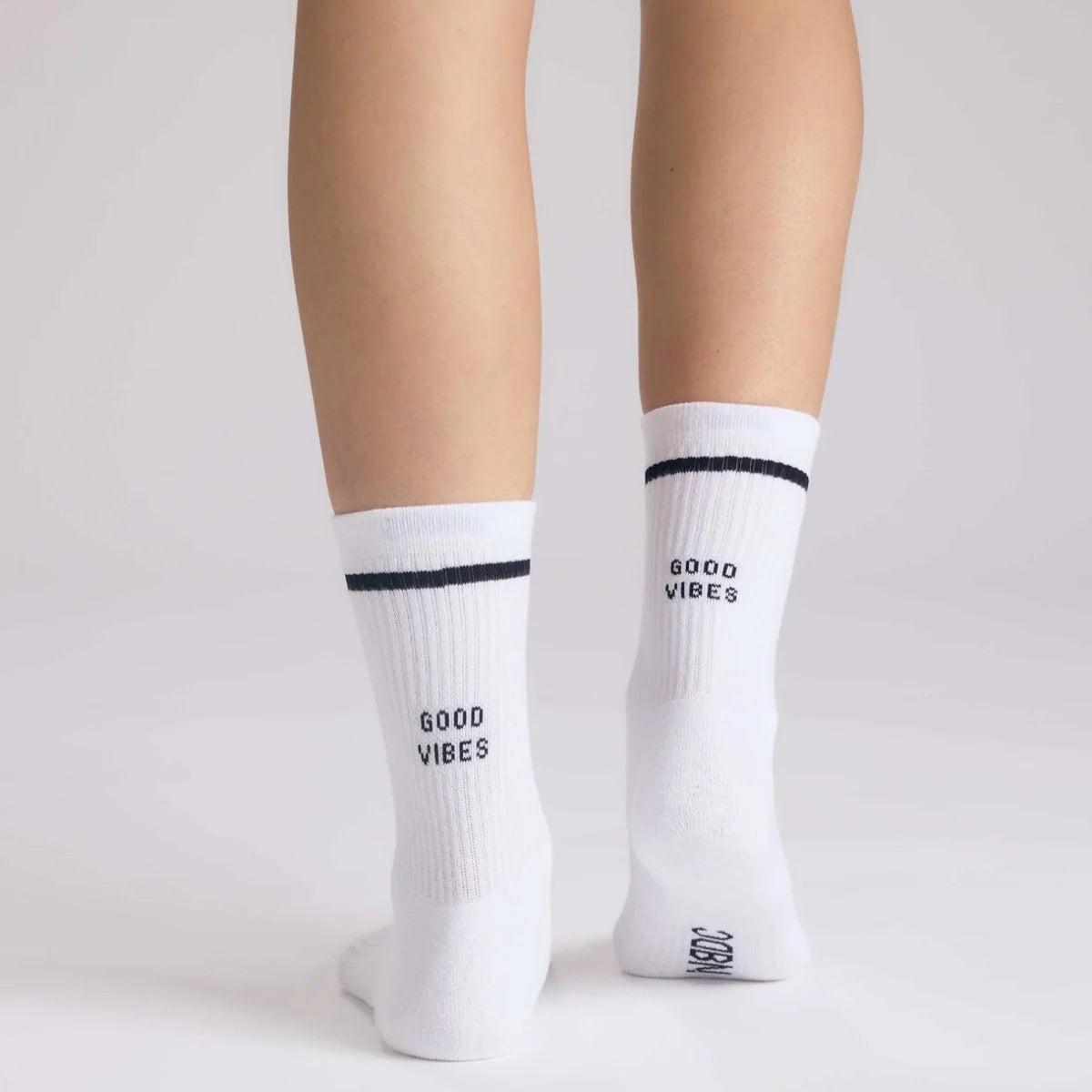 Eine Person, die auf einer weißen Fläche steht, von hinten gesehen, trägt weiße Socken mit einem schwarzen Streifen am oberen Ende und der Aufschrift GOOD VIBES auf der Rückseite jeder Socke.