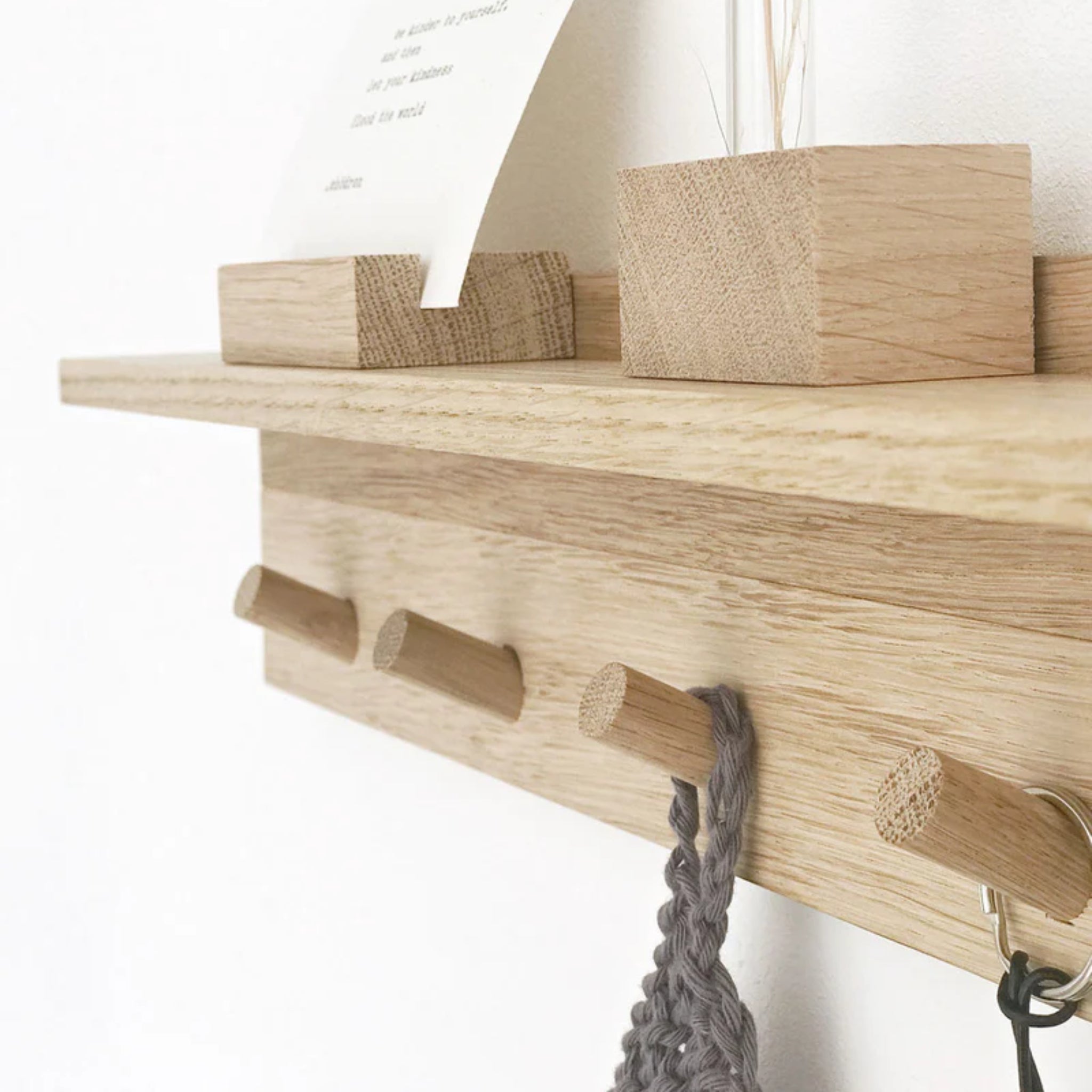 Nahaufnahme eines hellen Wandregals aus Holz mit drei runden Haken. An den Haken hängen ein graues Strickstück und ein Schlüsselanhänger, während oben auf dem Regal dekorative Holzklötze und eine Karte liegen.