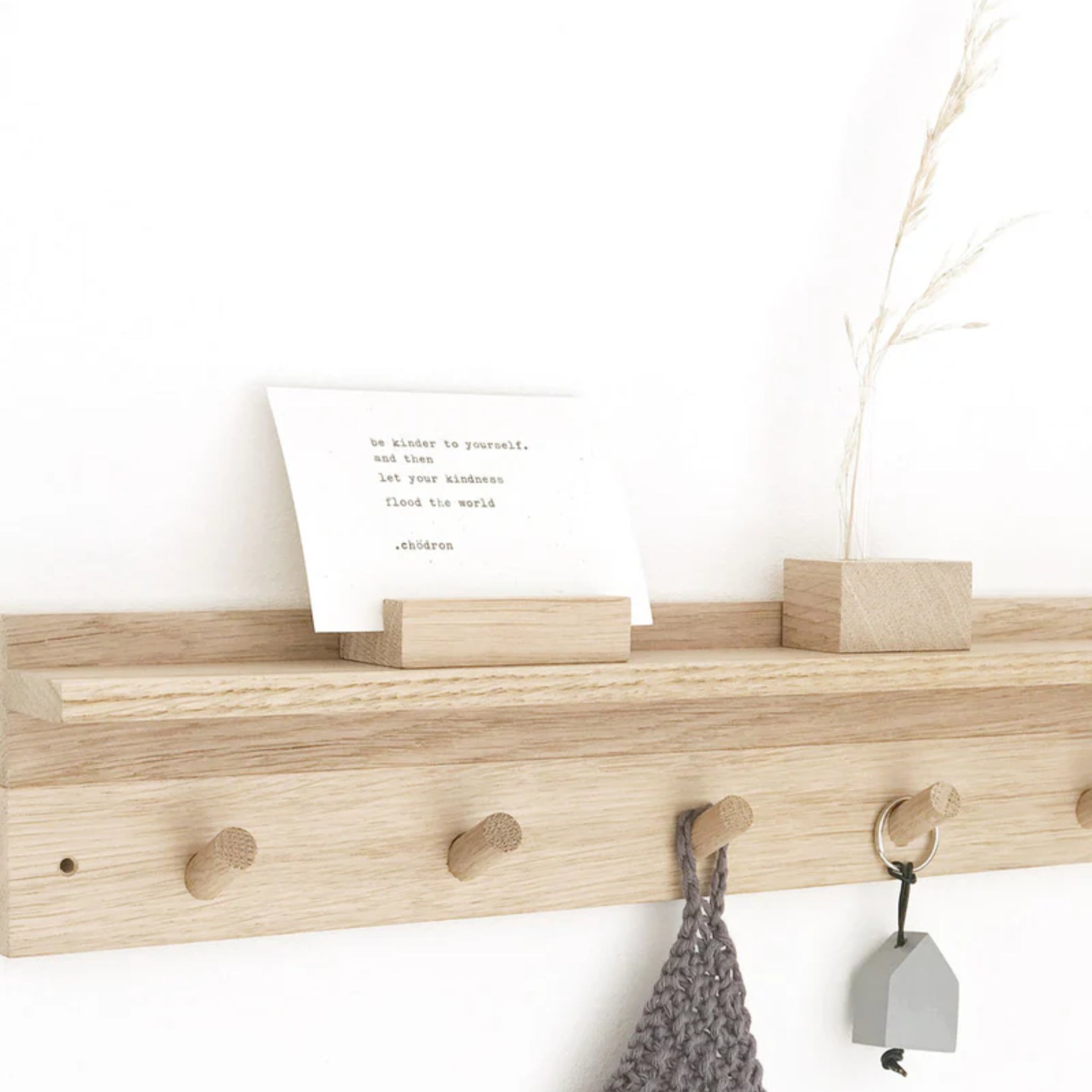 Ein helles Wandregal aus Holz mit fünf Stiften hält ein graues Strickteil und einen Schlüssel mit einem hausförmigen Schlüsselanhänger. Oben befindet sich ein Schlitz mit einem Zettel und eine kleine Holzvase mit getrocknetem Gras.