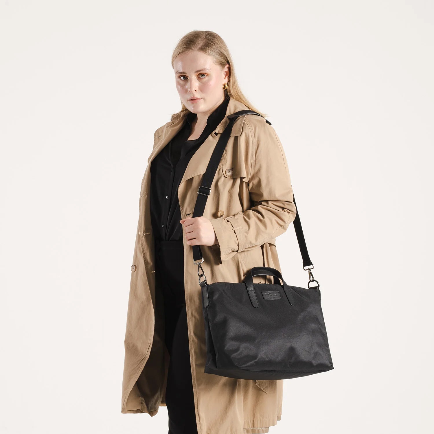 Eine Frau, die einen beigen Trenchcoat über schwarzer Kleidung trägt, hält eine schwarze Umhängetasche mit kurzen Griffen und einem langen Riemen vor einem schlichten weißen Hintergrund.