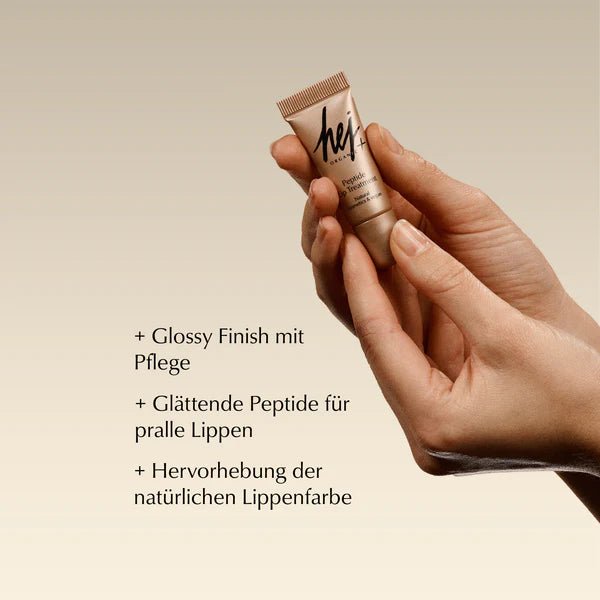 Eine Hand hält eine kleine Tube eines Lippenprodukts mit der Aufschrift "heji". Der Text auf Deutsch auf der linken Seite listet die Vorteile auf: glänzendes Finish mit Pflege, glättende Peptide für vollere Lippen und Verstärkung der natürlichen Lippenfarbe.