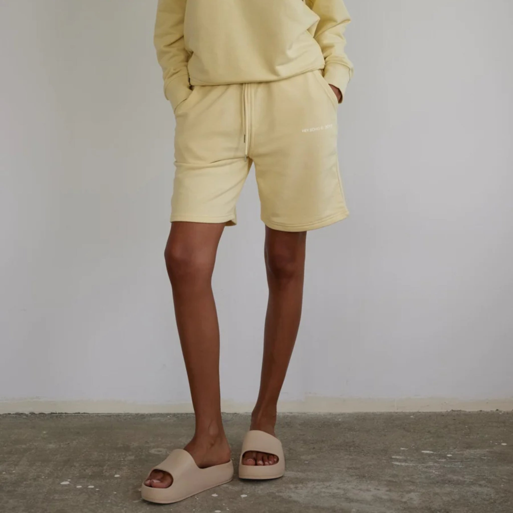 Person in gelbem Sweatshirt und Shorts mit hellbraunen Sandalen, steht auf grauem Betonboden vor weißer Wand.