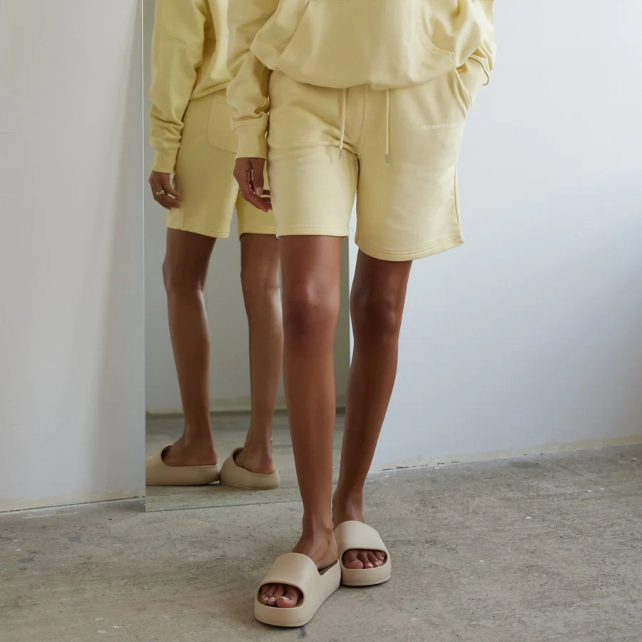 Person in gelbem Outfit und Sandalen steht vor einem Spiegel, der die Beine und den Boden reflektiert.