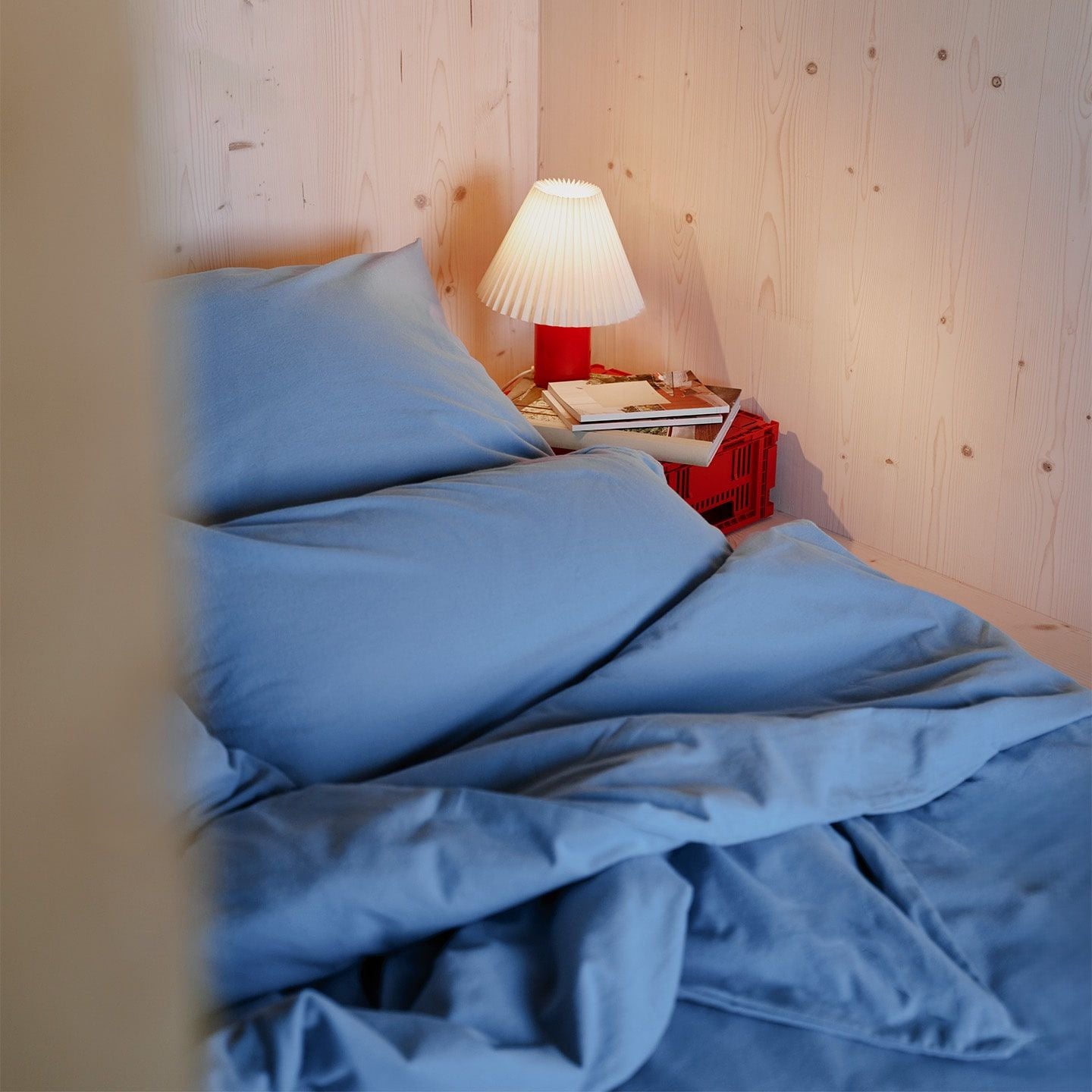 Ein Bett mit blauem Bettzeug steht neben einem Nachttisch mit Büchern und einer Lampe in einem holzverkleideten Raum.