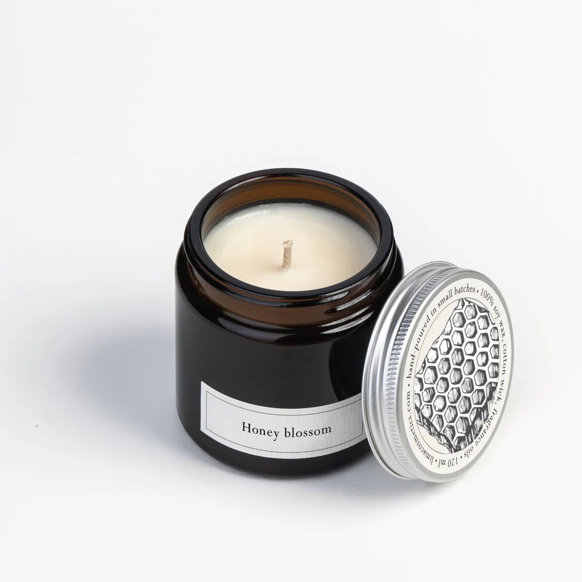 Eine braune Glaskerze mit der Aufschrift Honey blossom, mit cremig-weißem Wachs und einem einzelnen Docht, neben dem silbernen Deckel mit Wabenmuster.