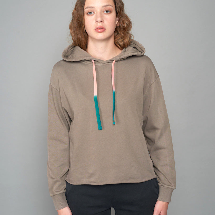 Frau in grauen Kapuzenpullover mit zweifarbigen Kordeln steht vor grauem Hintergrund.