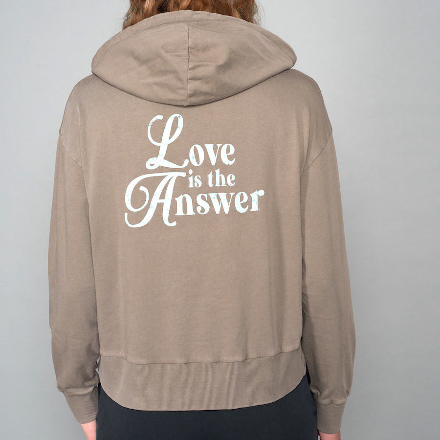 Person trägt einen braunen Kapuzenpullover mit dem weißen Schriftzug "Love is the Answer" auf dem Rücken.
