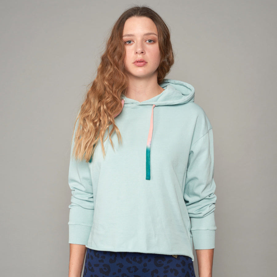 Frau mit langen Haaren trägt einen hellblauen Hoodie mit zweifarbigen Kordeln und dunkelblauer Hose mit Muster.