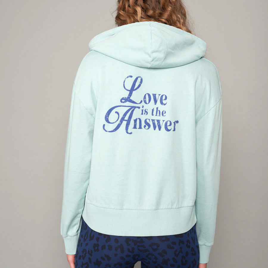 Person mit mintgrünem Hoodie, auf dem Rücken "Love is the Answer" steht, trägt gemusterte blaue Leggings.