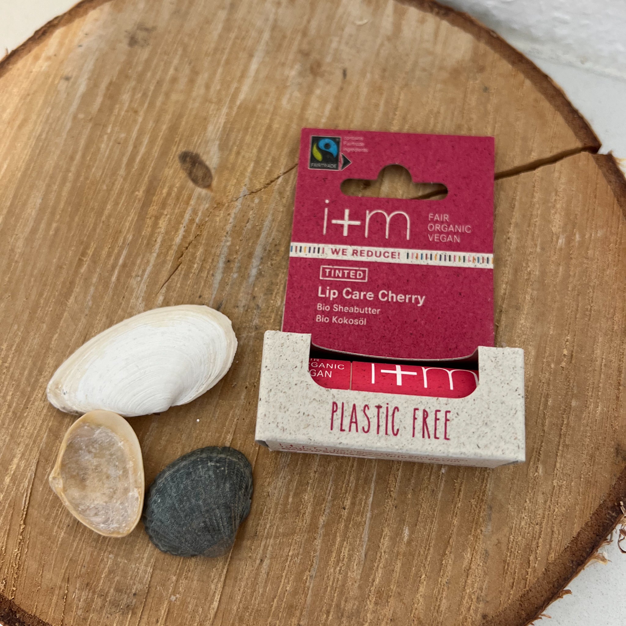 Eine Verpackung von rotem Lip Balm mit der Aufschrift "i+m", neben drei Muscheln auf Holz.