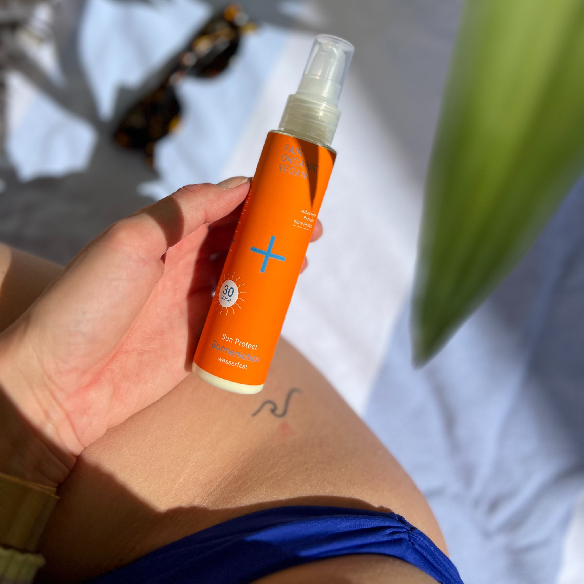 Eine Hand hält eine orangefarbene Flasche Sonnenschutzlotion mit LSF 30, im Hintergrund eine helle Decke.
