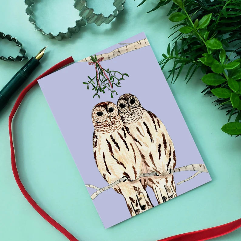 Grußkarte mit zwei Eulen auf einem Ast, zusammen unter Mistelzweigen, umgeben von Pflanzen und Backformen.