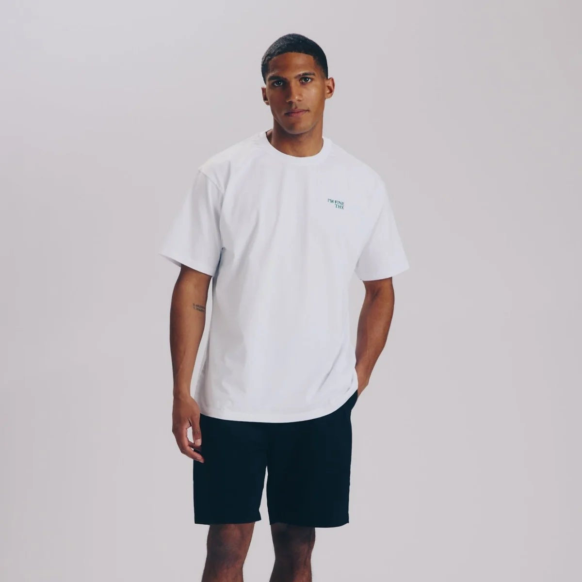 Ein Mann steht vor einem schlichten hellgrauen Hintergrund und trägt ein weißes T-Shirt, schwarze Shorts, weiße Socken mit schwarzen Streifen und weiße Turnschuhe. Eine Hand steckt in seiner Tasche und er hat einen neutralen Gesichtsausdruck.