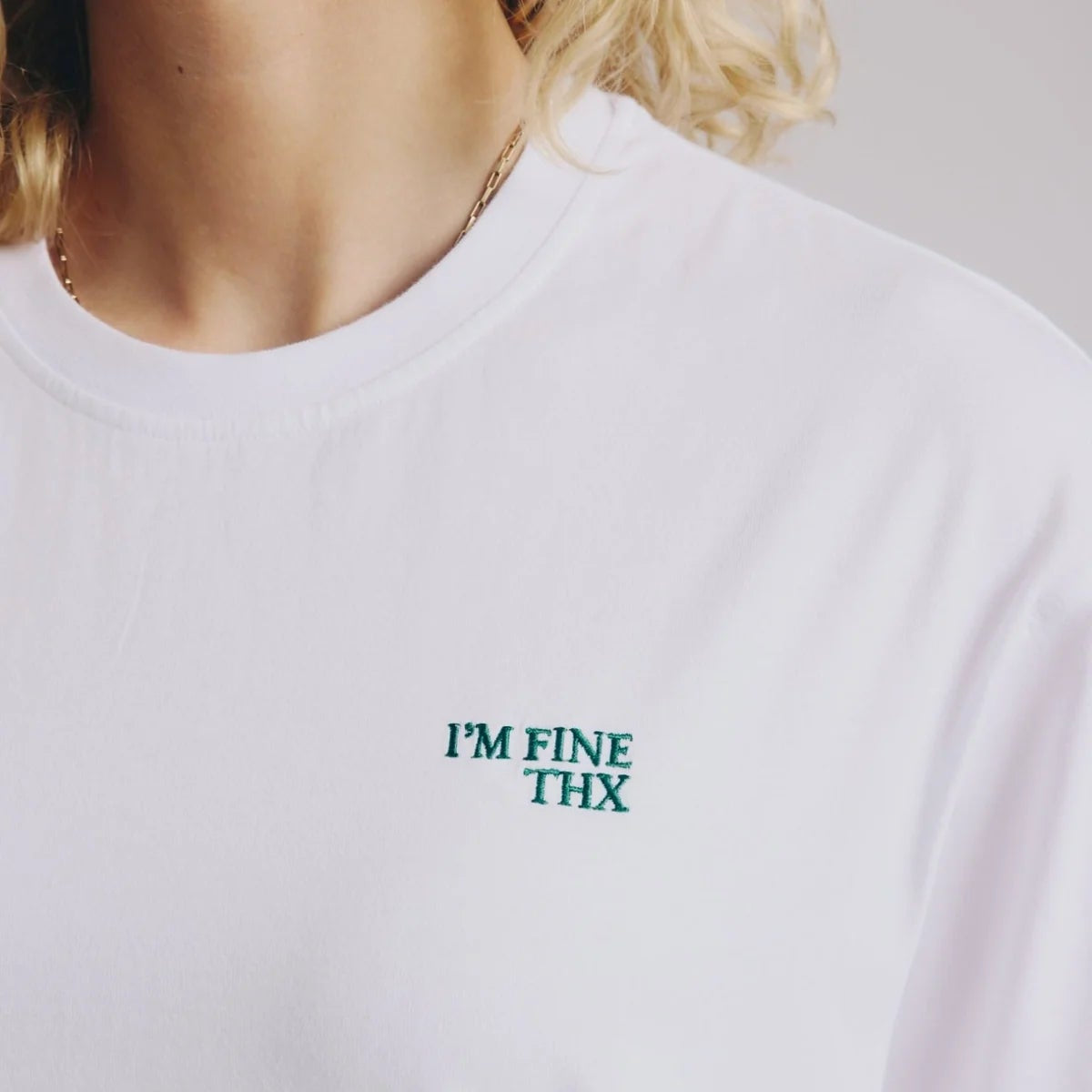Eine Person trägt ein schlichtes weißes T-Shirt mit der Aufschrift IM FINE THX in grüner Schrift auf der linken Seite der Brust. Das Gesicht der Person ist nicht zu sehen.