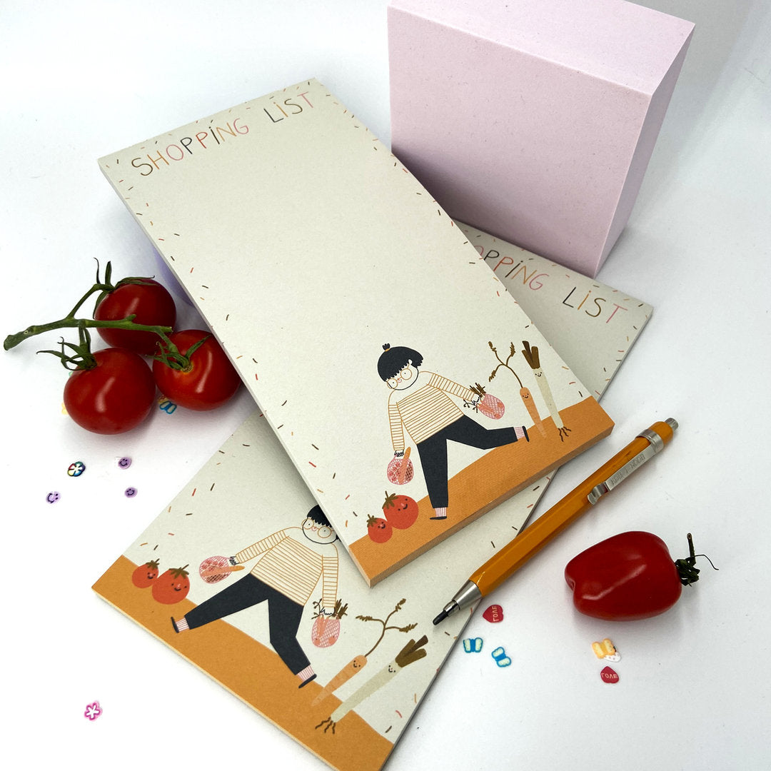 Notizblöcke mit "Shopping List" und Illustrationen, umgeben von Tomaten, bunten Perlen und einem Stift.