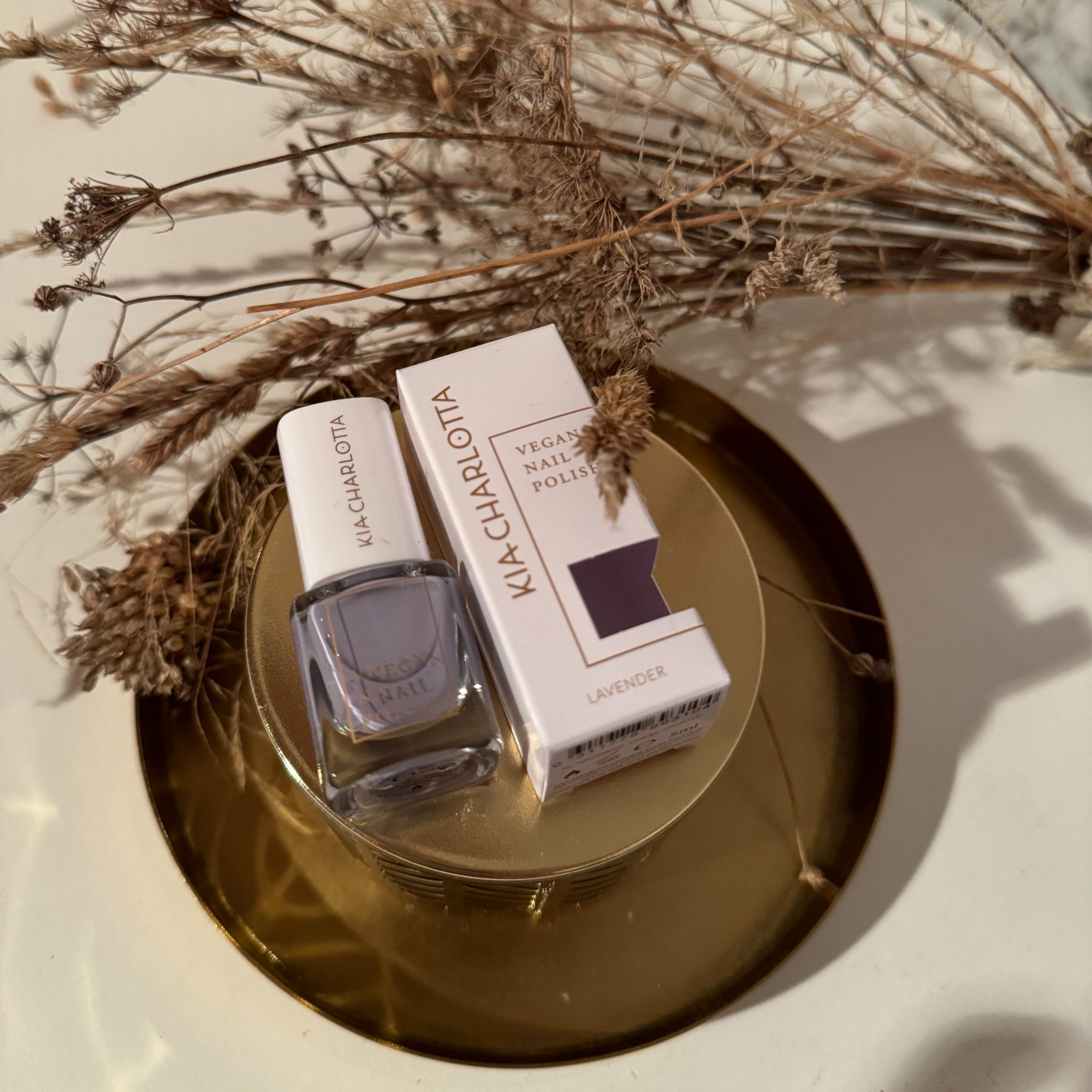 Nagellackflasche und -verpackung auf einem goldenen Tablett mit getrockneten Pflanzen.