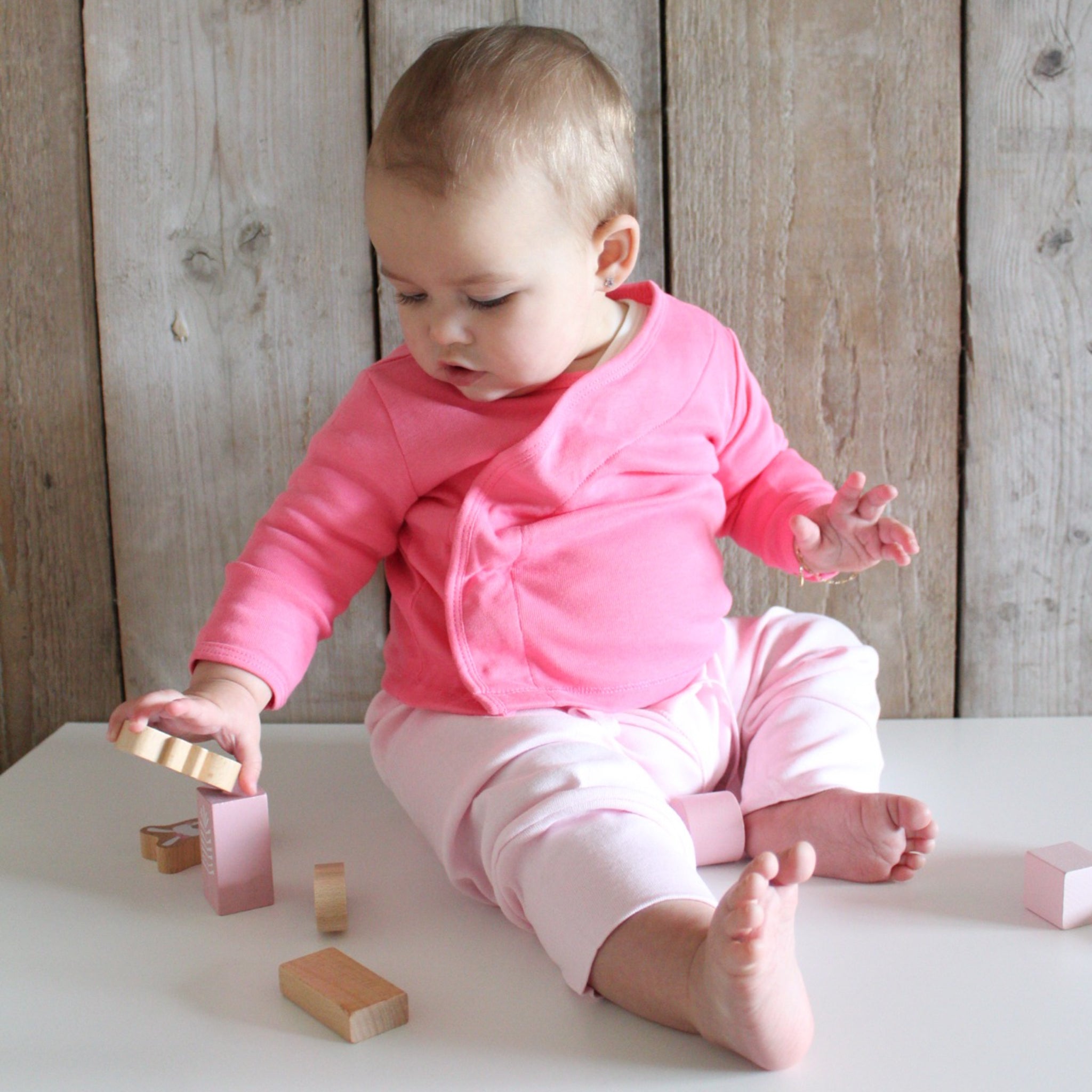 Ein Baby in rosa Kleidung spielt mit Holzklötzen auf einem weißen Tisch vor einem Holzhintergrund.