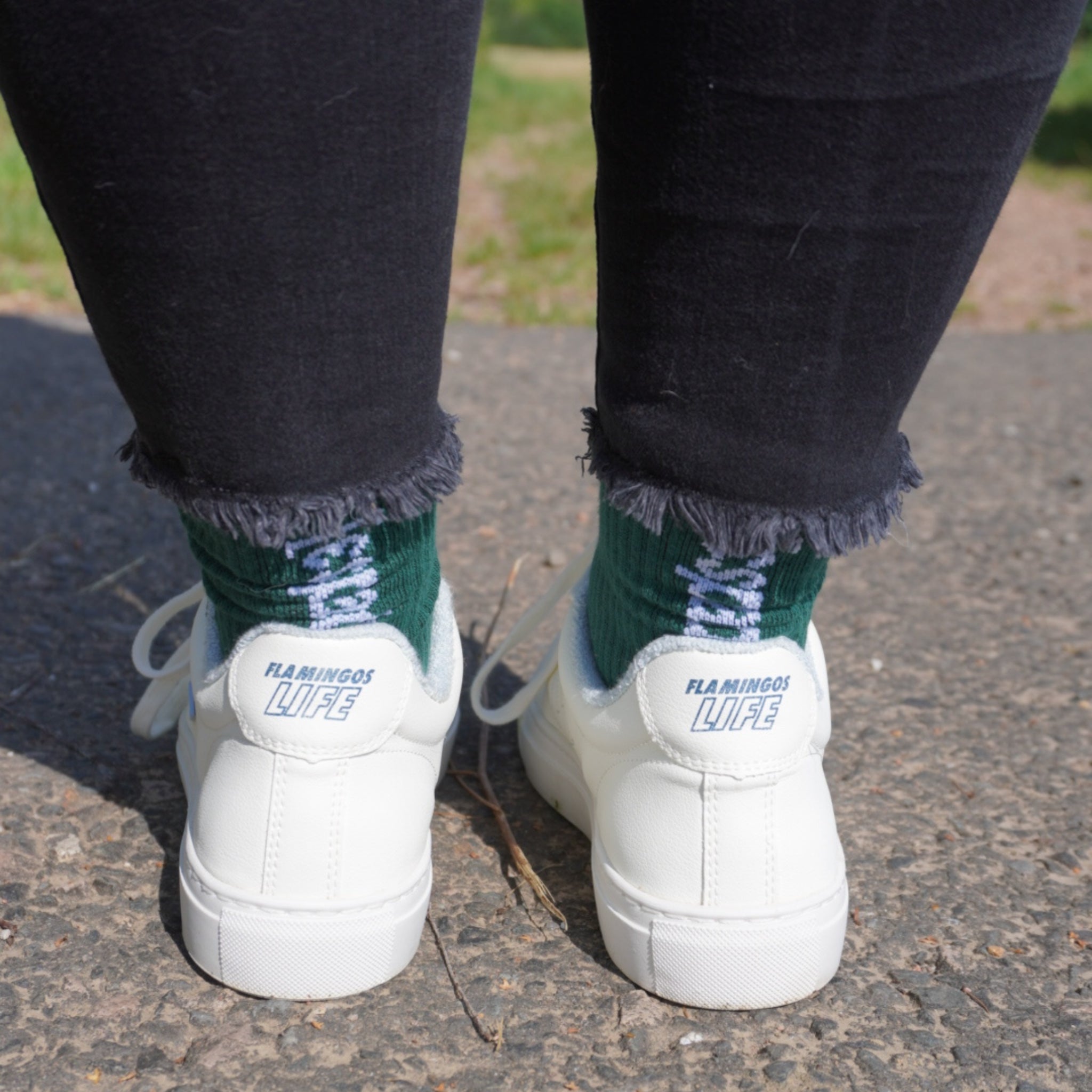 Person trägt weiße Sneaker und grüne Socken mit Fransenjeans auf einem gepflasterten Weg.