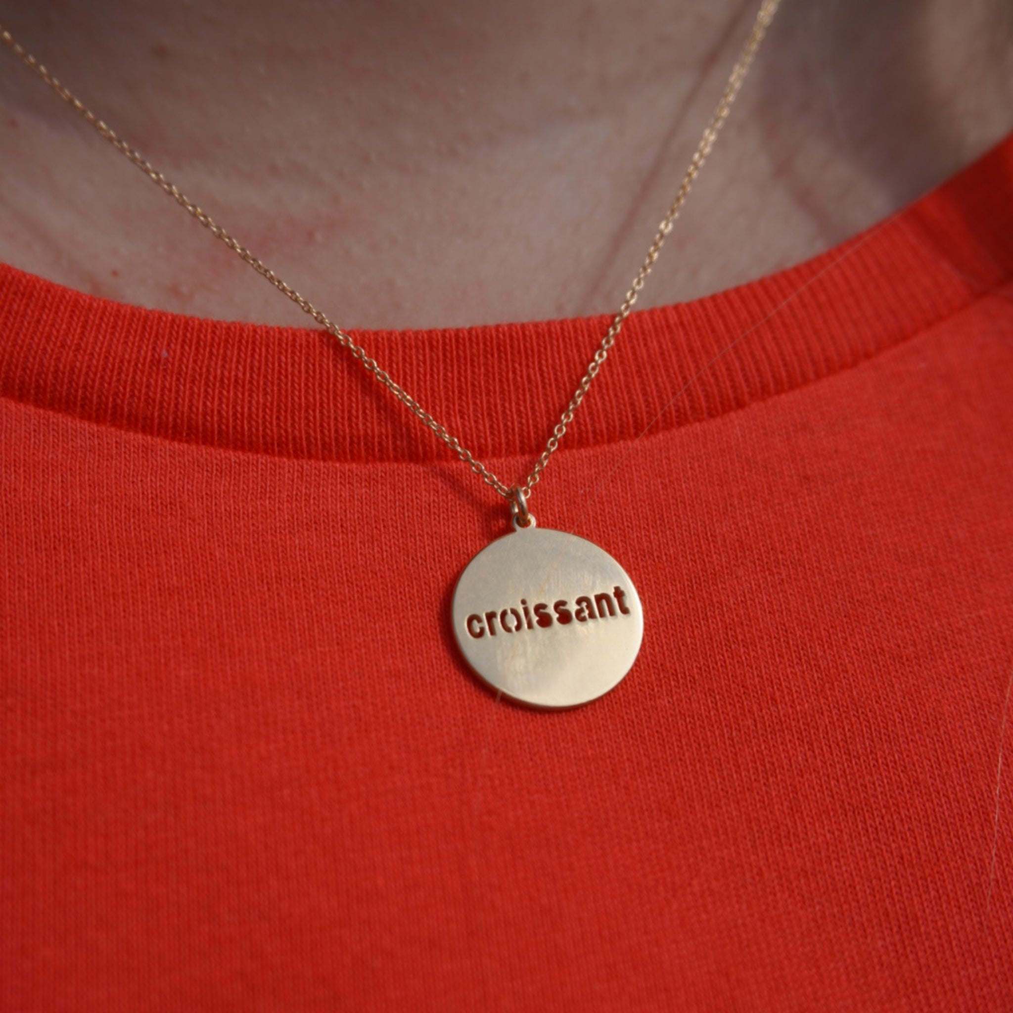 Goldene Halskette mit rundem Anhänger auf rotem Oberteil, Anhänger trägt die Gravur "croissant".