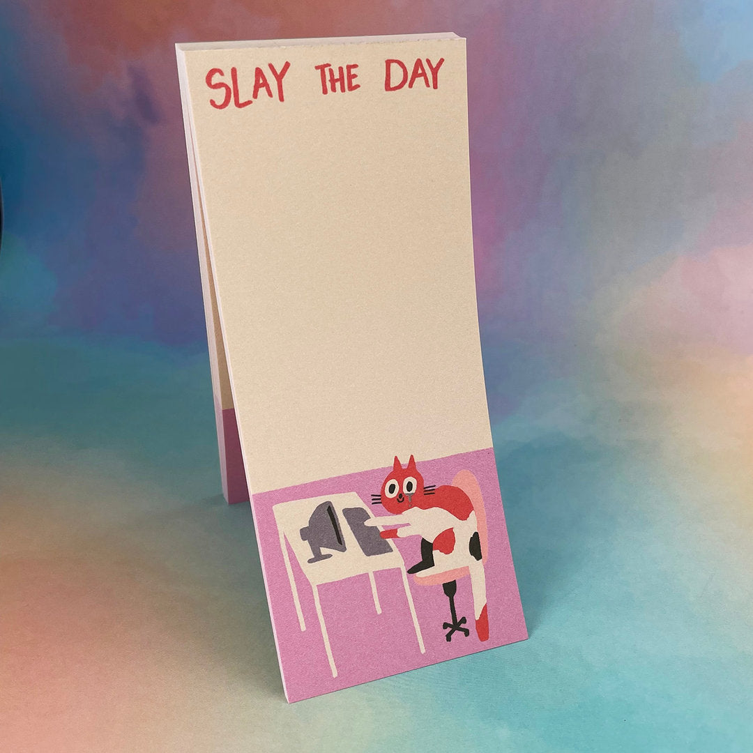 Notizblock mit der Aufschrift "Slay the Day", darunter eine cartoonartige Katze an einem Computer auf einem lilafarbenen Hintergrund.