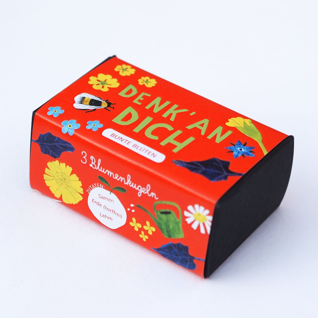 Eine kleine, rechteckige rote Schachtel mit der Aufschrift "DENK' AN DICH" mit illustrierten Blumen, einer Biene und einer Gießkanne. Die Schachtel enthält 3 Blumensaatkugeln und hat Blumenverzierungen auf der Verpackung.