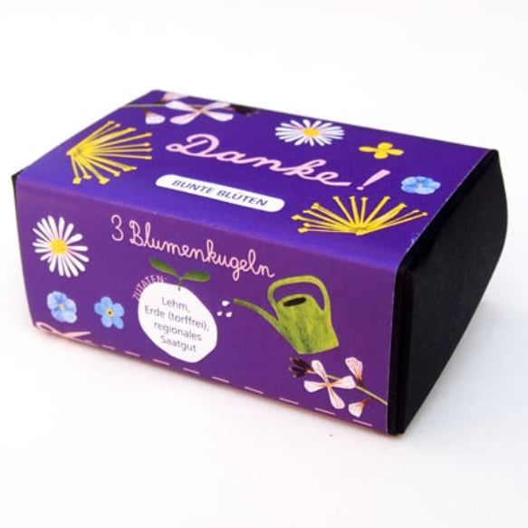 Eine kleine lila Schachtel mit Blumenillustrationen und einer grünen Gießkanne. Auf der Schachtel steht "Danke", "3 Blumenkugeln" und "Bunte Blüten" auf Deutsch, was bedeutet, dass sie Blumenkugeln enthält.
