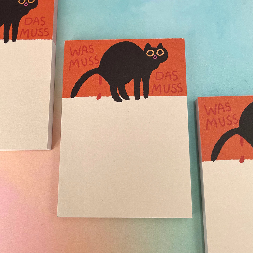 Notizblöcke mit einem schwarzen Katzenmotiv und der Aufschrift "WAS MUSS DAS MUSS" auf orangem Hintergrund.