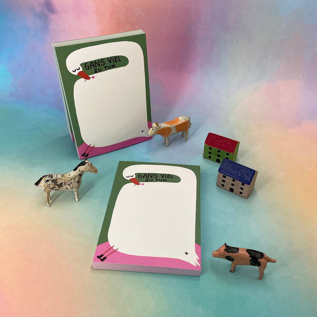 Notizblöcke mit "Gans viel zu tun"-Aufschrift, umgeben von Spielzeugtieren und Häusern auf buntem Hintergrund.