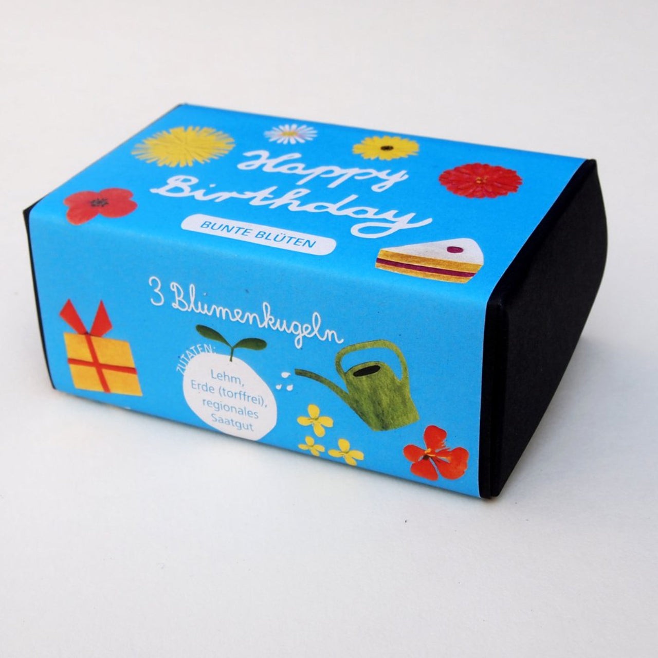 Eine kleine blaue Schachtel, verziert mit bunten Zeichnungen von Blumen, einem Stück Kuchen, einem Geschenk und einer Gießkanne. Auf der Schachtel steht "Happy Birthday" und "3 Blumenkugeln" auf Deutsch, mit zusätzlichem Text und Illustrationen.