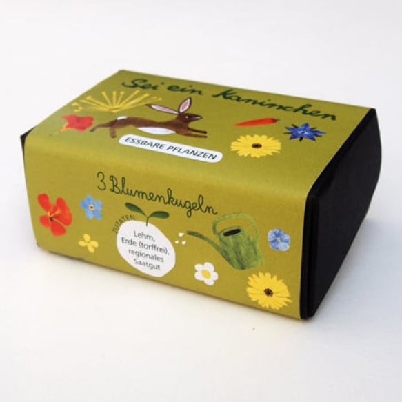 Eine kleine grün-schwarze Schachtel, verziert mit illustrierten Blumen, einem Kaninchen und deutschem Text, darunter "3 Blumenkugeln" und "Essbare Pflanzen", auf weißem Hintergrund.