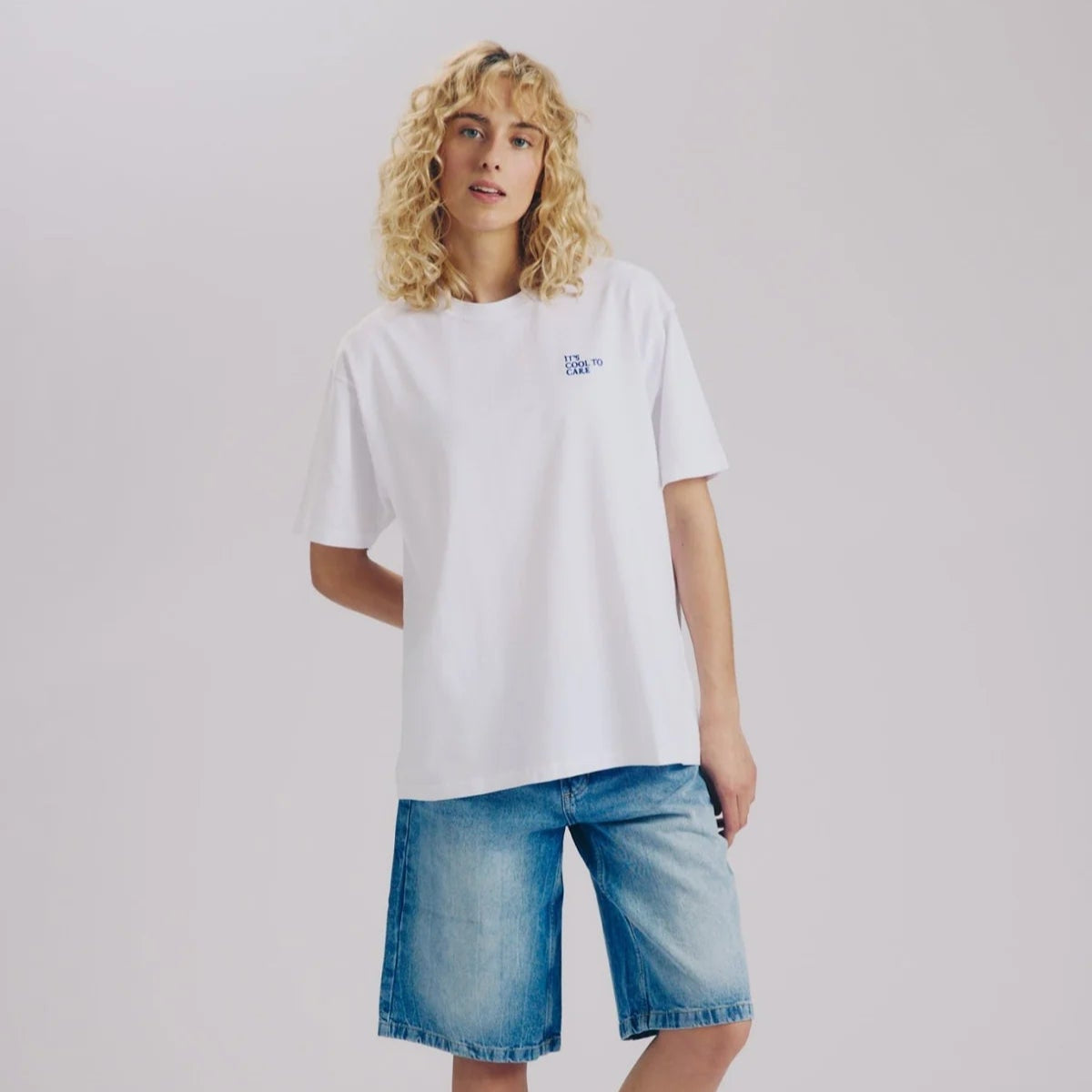 Eine Person mit lockigem blondem Haar trägt ein lockeres weißes T-Shirt, knielange blaue Jeans-Shorts, weiße Socken und weiße Turnschuhe und steht vor einem schlichten hellen Hintergrund.