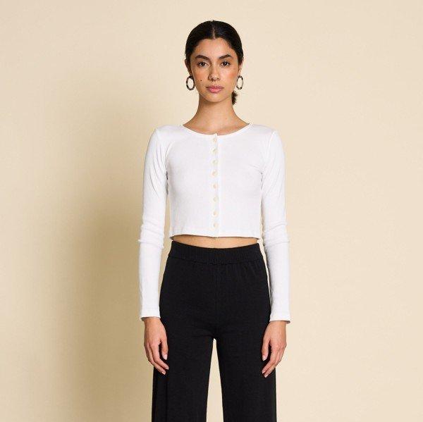 Frau in weißem langärmligen Oberteil und schwarzen Hosen vor beigem Hintergrund.