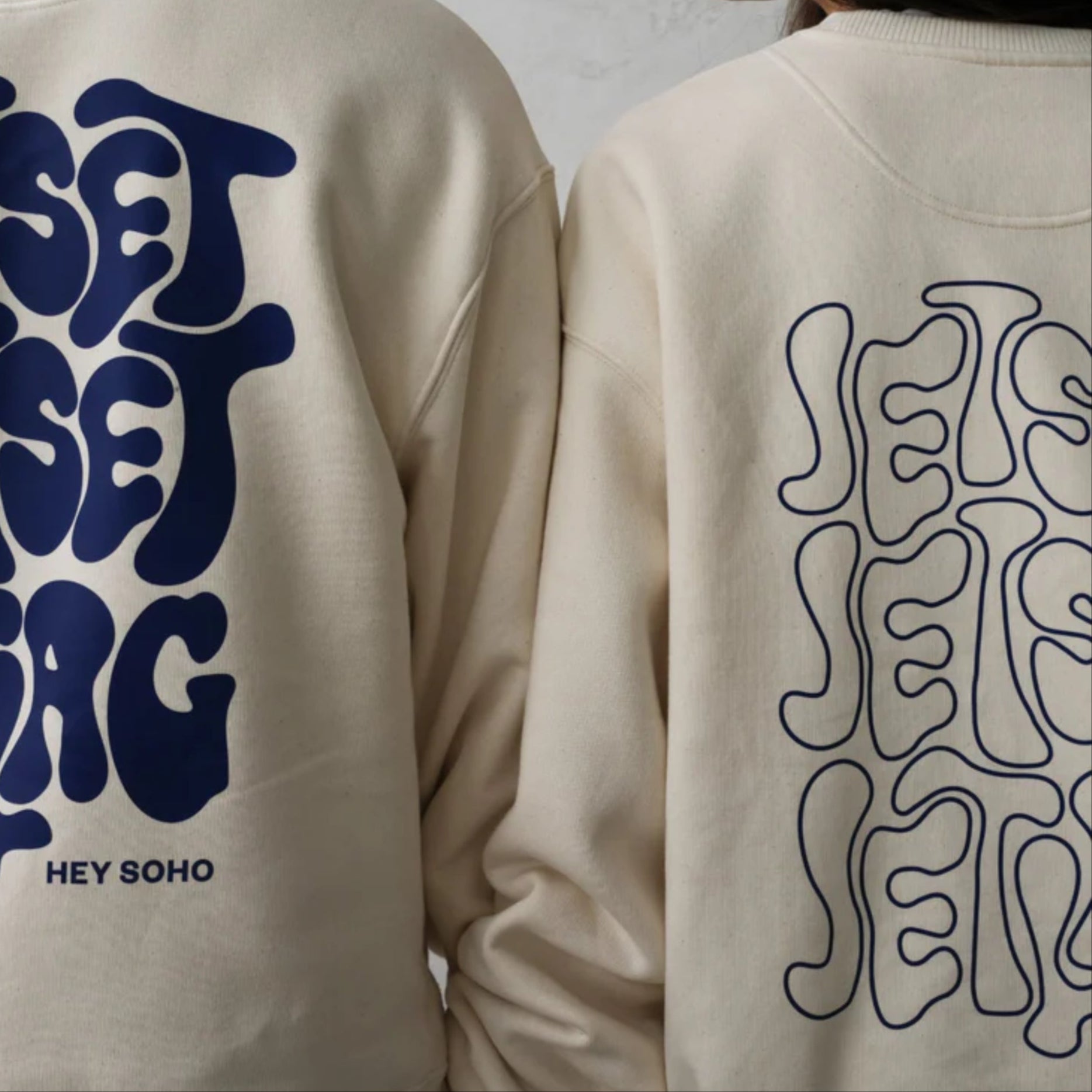Zwei Personen tragen beige Sweatshirts mit blauen, verspielten Schriftzügen "Jet Set" und "Hey Soho" auf der Rückseite.