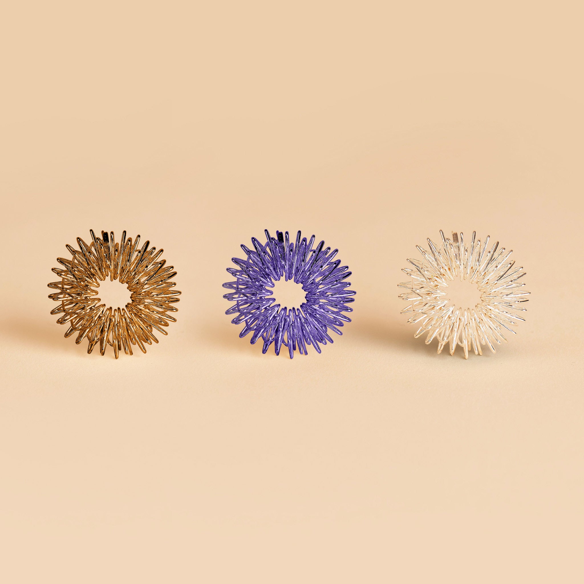 Drei spiralförmige Massage-Ringe in Gold, Lila und Silber auf beigem Hintergrund.