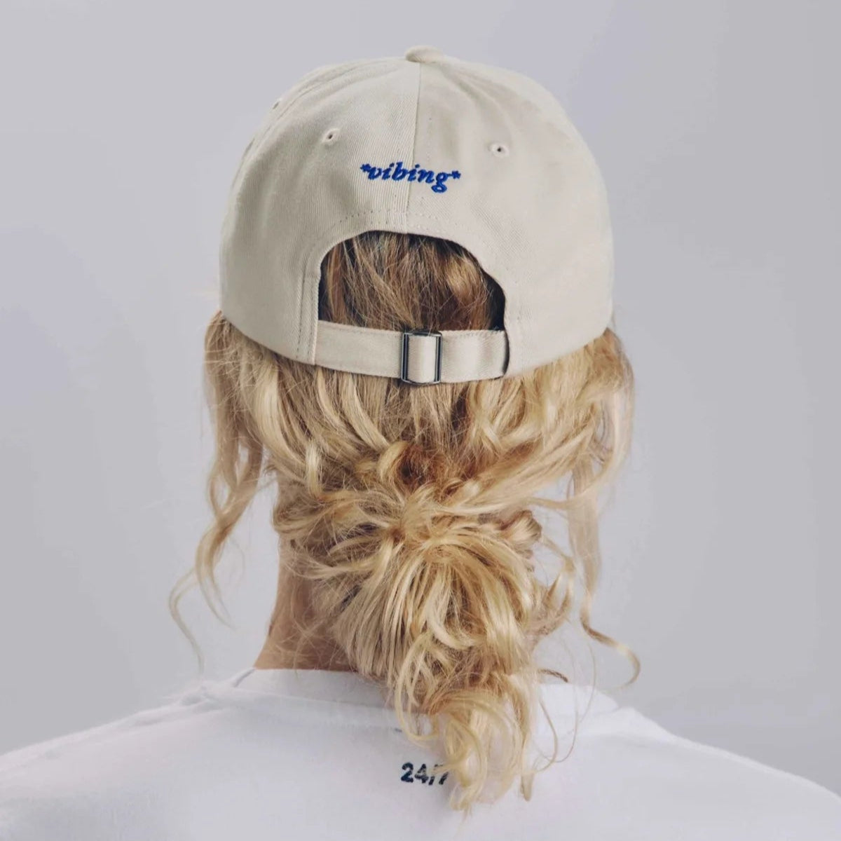 Eine Person mit blonden, gelockten Haaren, die zu einem niedrigen, unordentlichen Dutt gebunden sind, trägt eine hellbeige Baseballmütze mit dem blau aufgestickten Wort Vibing auf dem Rücken und ein weißes T-Shirt. Das Foto ist von hinten aufgenommen.