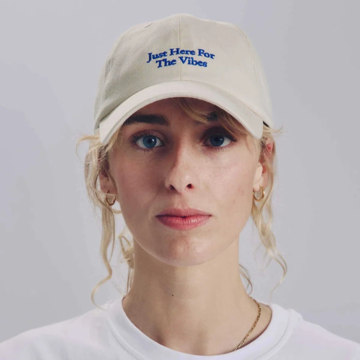Eine Person mit hellem Haar trägt eine cremefarbene Baseballkappe mit der Aufschrift Just Here For The Vibes in blauer Schrift und ein schlichtes weißes T-Shirt vor einem schlichten hellen Hintergrund.