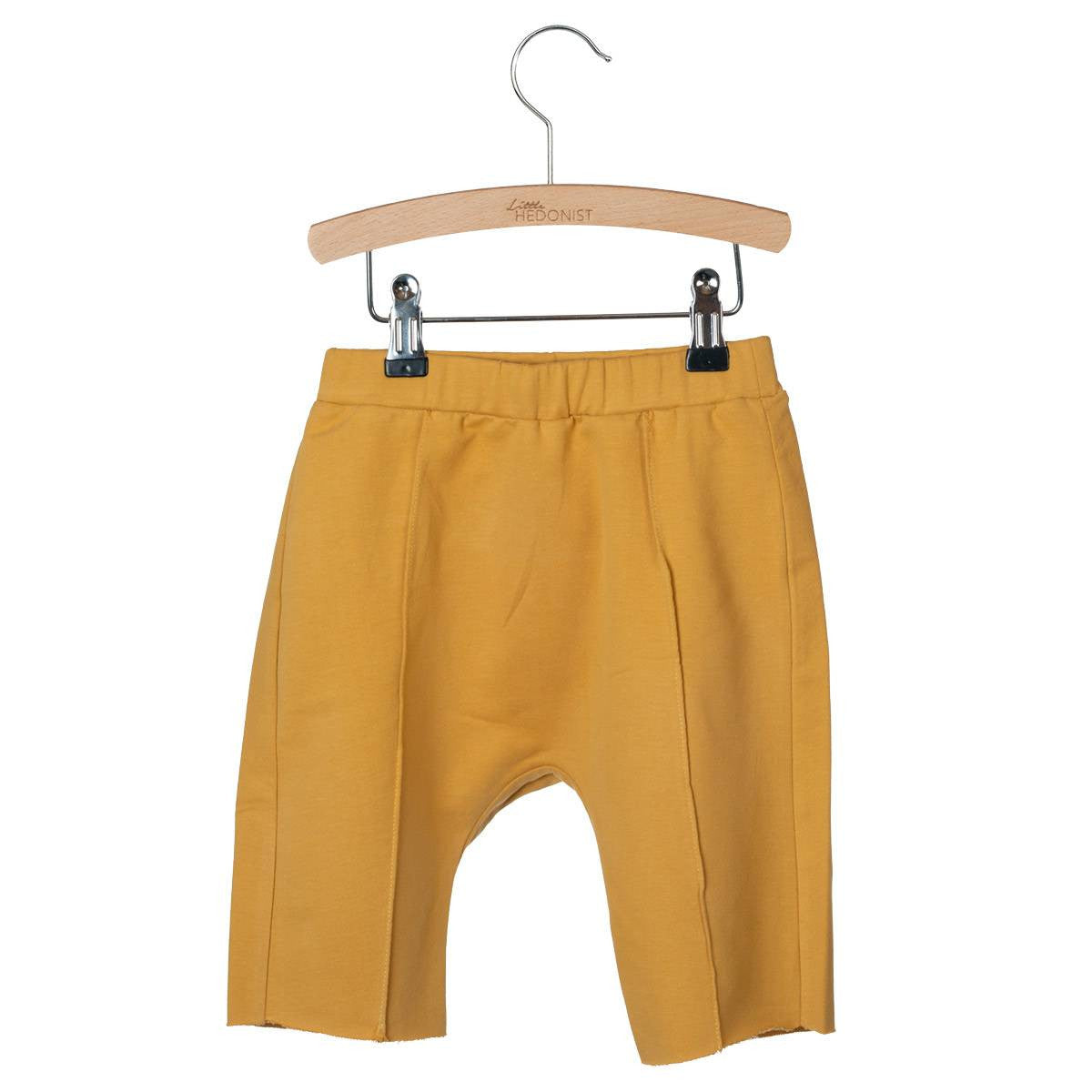 Eine gelbe Hose für Kleinkinder mit einem elastischen Bund hängt an einem Holzbügel mit Metallklammern vor einem weißen Hintergrund.