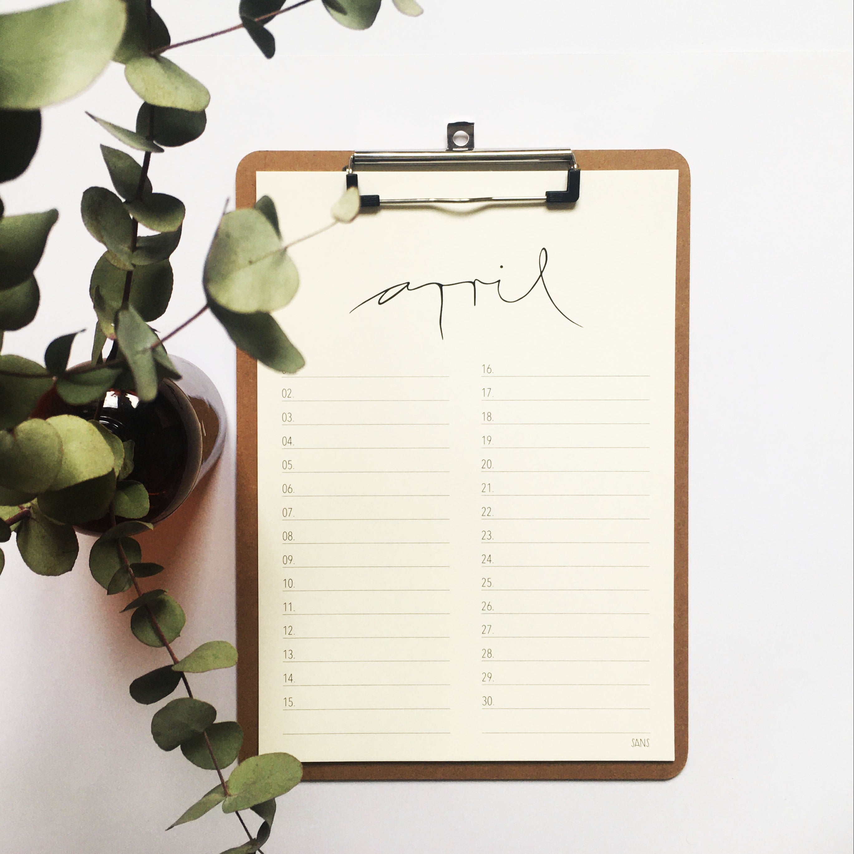 Ein Klemmbrett mit einem minimalen Kalenderblatt für April mit handgeschriebenem Text, neben einer Topfpflanze mit grünen Blättern auf weißem Hintergrund.