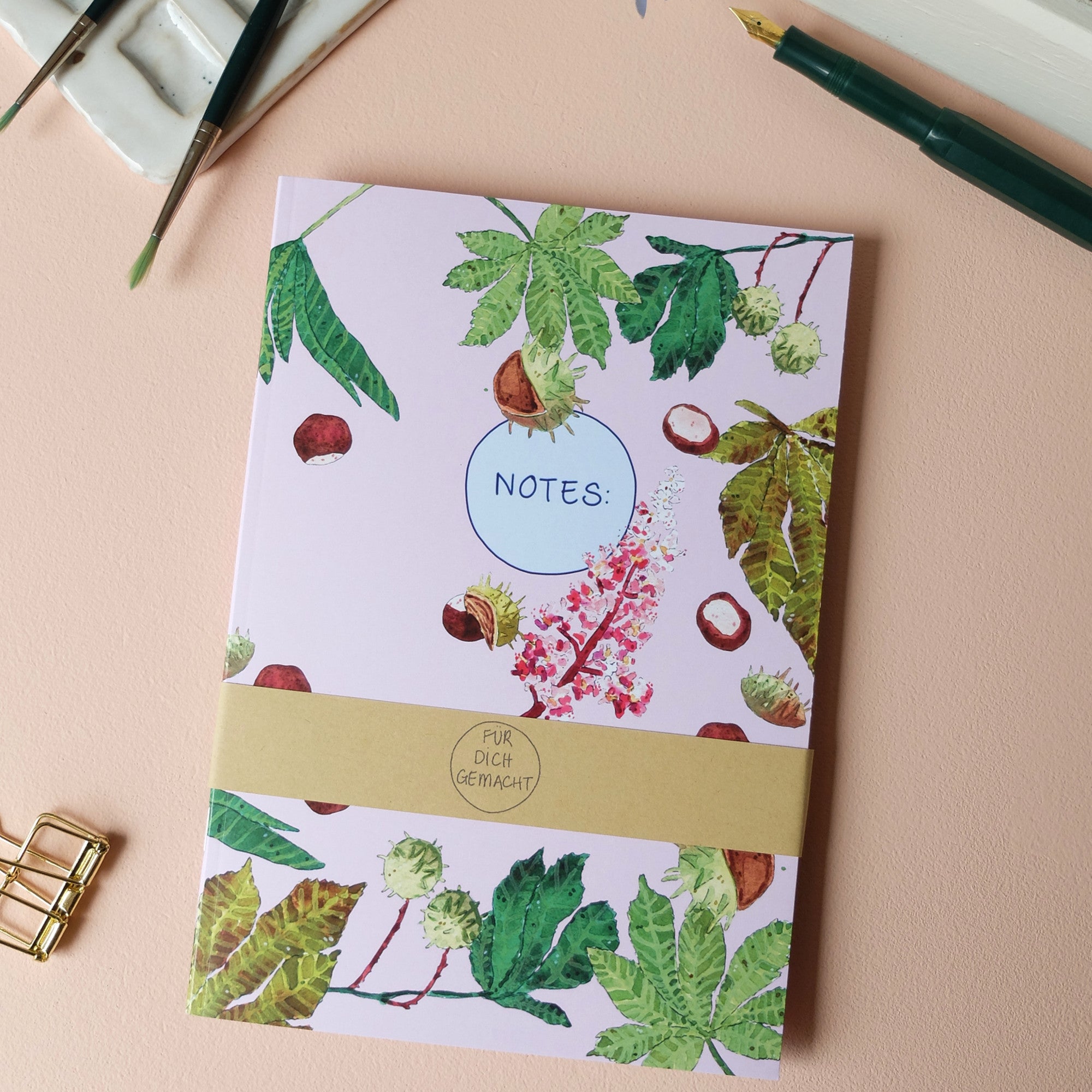 Notizbuch mit Kastanien- und Blätterdesign, umgeben von Malutensilien auf einem rosa Hintergrund.