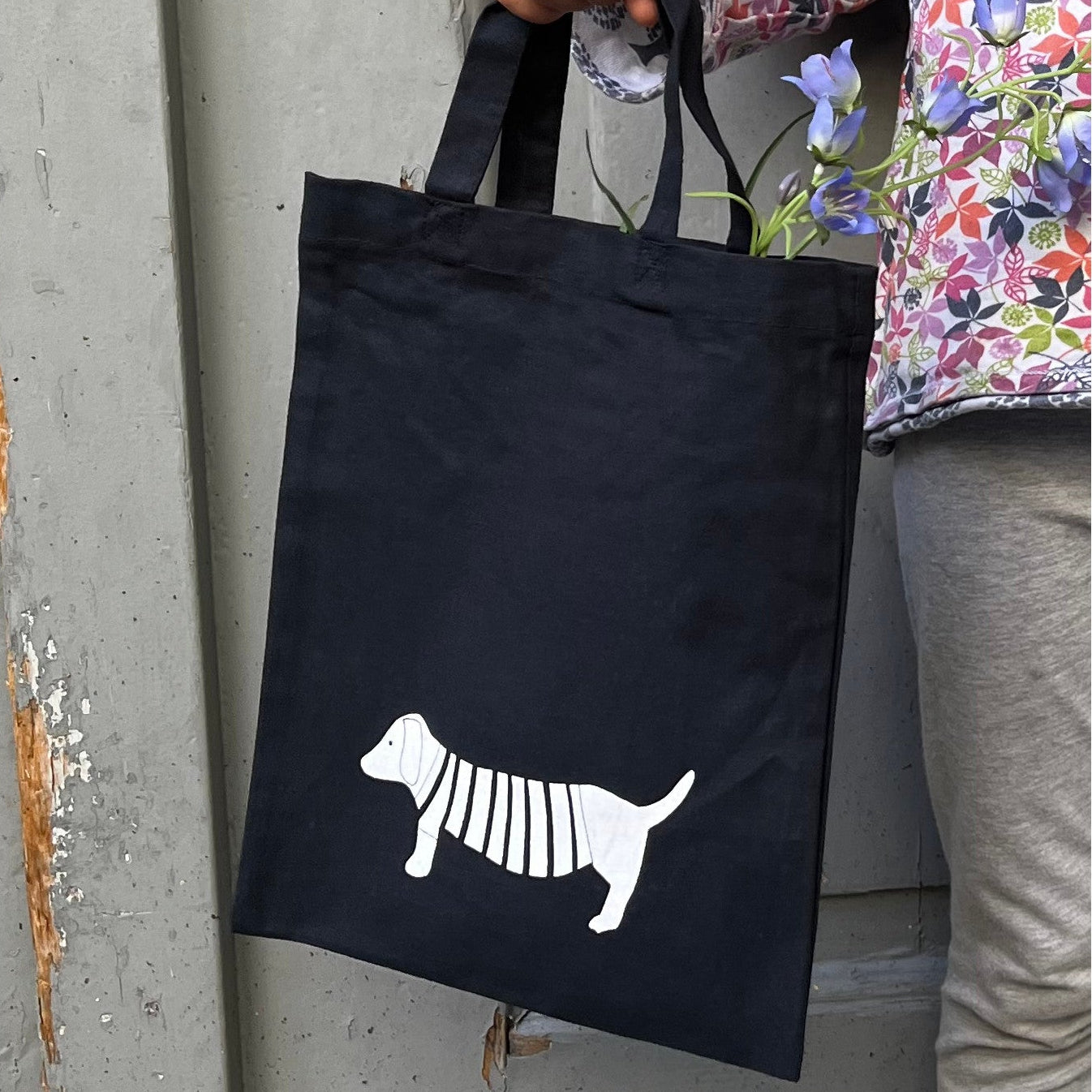 Eine Person hält eine schwarze Tragetasche, auf der ein weißer Hund abgebildet ist. Der Hund hat Streifen auf seinem Körper. Die Person trägt ein bunt geblümtes Hemd und eine graue Hose, aus der lila Blumen herausschauen.