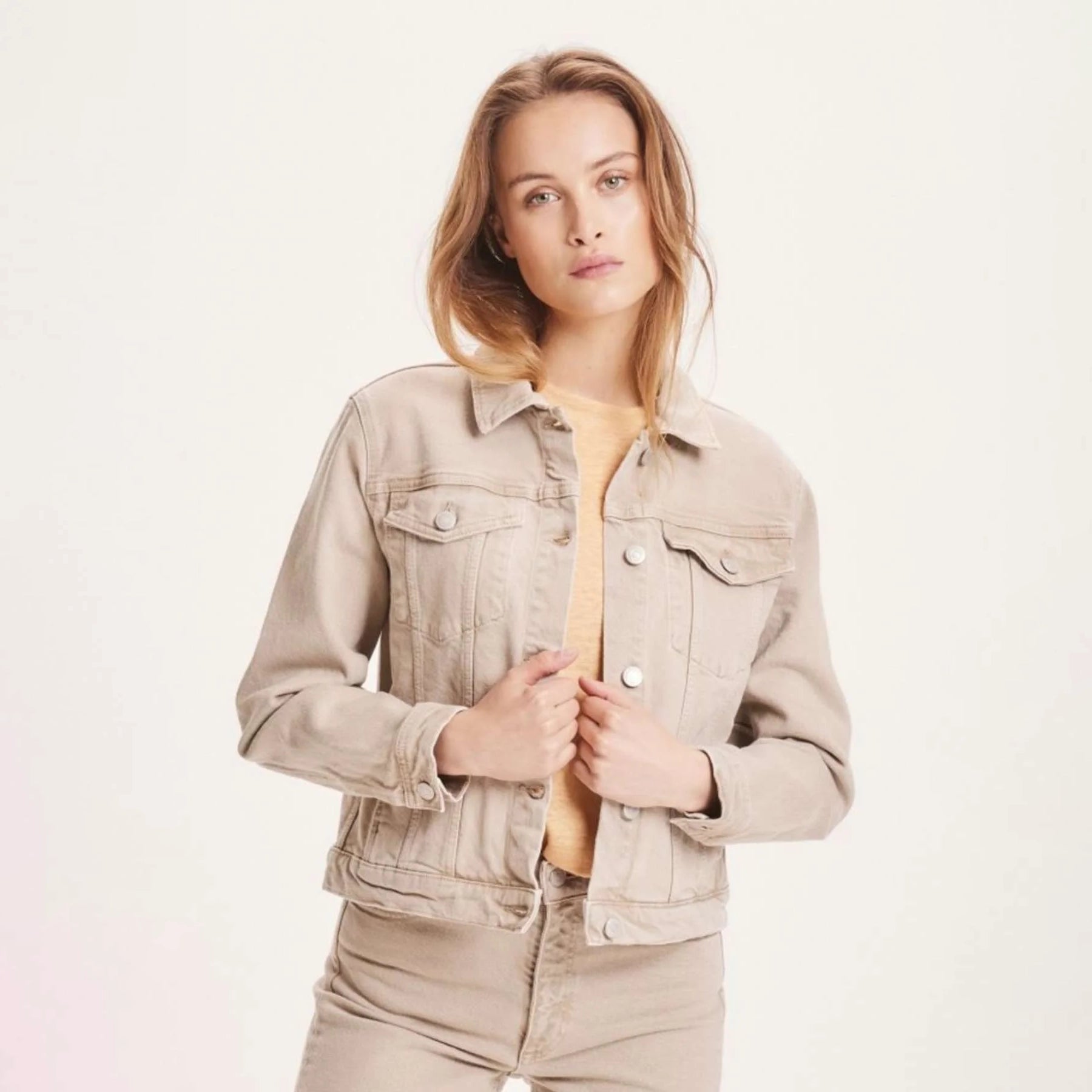 Frau in beiger Jeansjacke und Hose vor hellem Hintergrund.