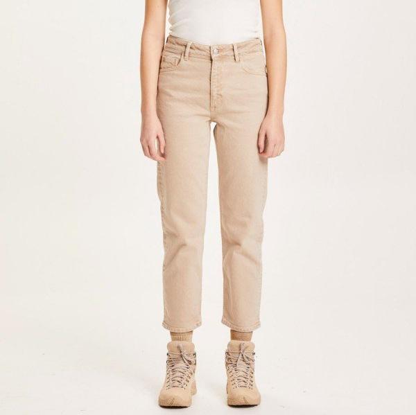 Person in cremefarbenen Jeans und beigem Schuhwerk, vor hellem Hintergrund stehend.