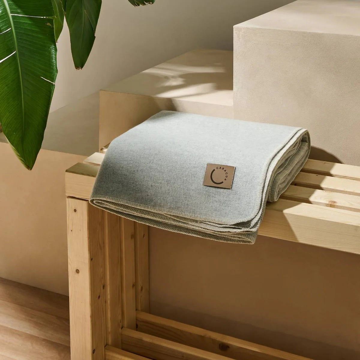 Eine ordentlich gefaltete hellgraue Decke mit einem kleinen Etikett liegt auf einer Holzbank; ein großes grünes Pflanzenblatt hängt von der linken Seite herab, und im Hintergrund befinden sich beigefarbene geometrische Flächen.