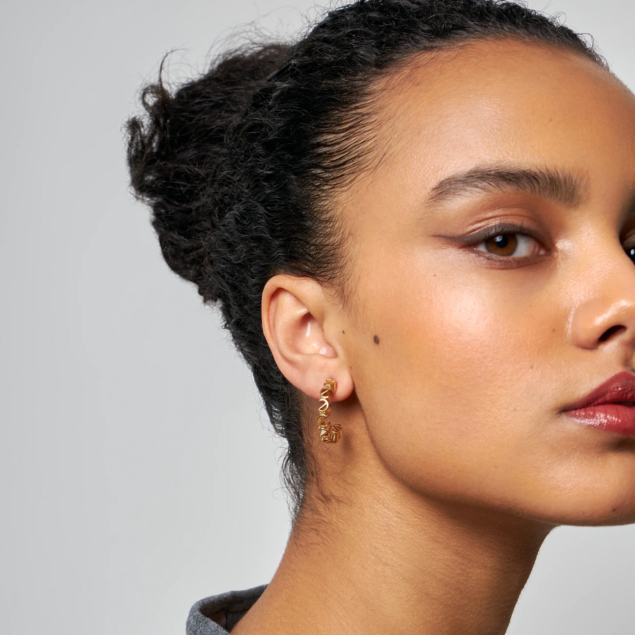 Nahaufnahme eines Gesichts mit hochgestecktem Haar, goldenen Ohrringen und natürlichem Make-up.