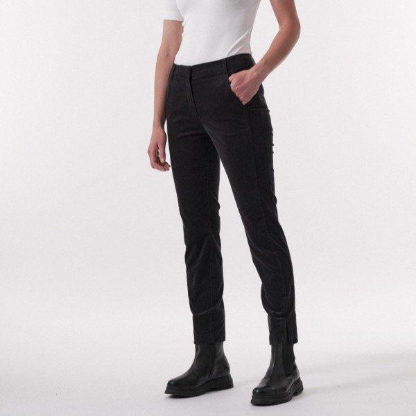 Person in schwarzer Hose, weißem Oberteil und schwarzen Stiefeln, posiert mit einer Hand in der Hosentasche auf neutralem Hintergrund.